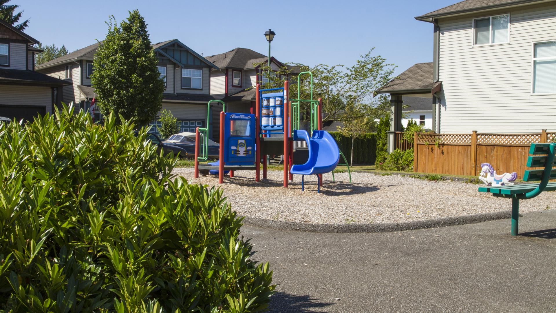 A paved park surrounded by houses. There is a small playground in the centre, on a bed of gravel.